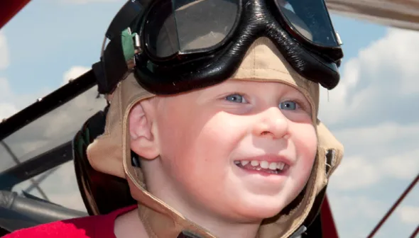 Un enfant souriant porte une chemise rouge, un casque d'aviateur beige et de grosses lunettes d'aviateur noires. Derrière lui, on aperçoit un ciel bleu nuageux et le sommet d'une aile de biplan rouge.