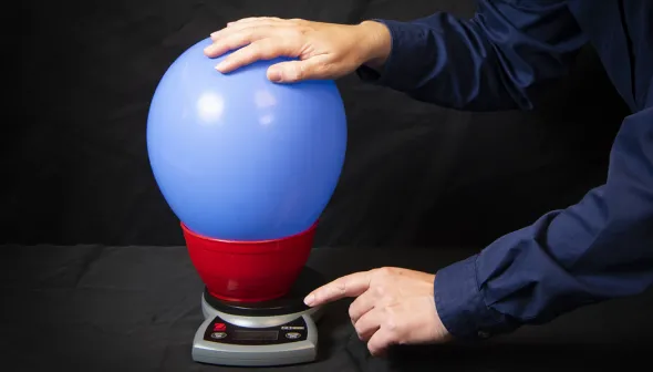 Close-up view of two hands holding and pointing at an inflated blue balloon which is sitting in a smaller red container.