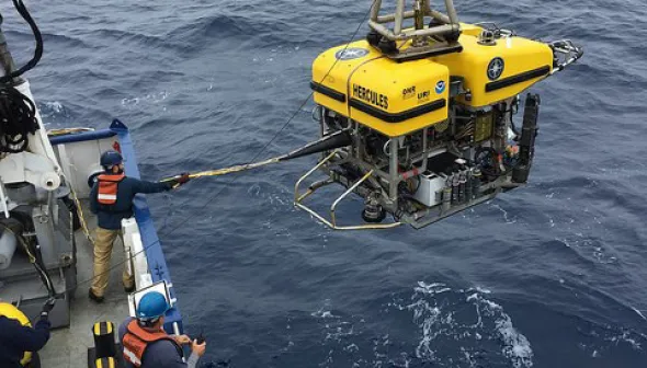A crane on a boat holds a remotely operated vehicle (ROV) over the water as it is deployed.