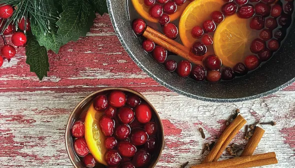 An aerial view shows a metal container and a cup filled with cider; red cranberries and orange slices are floating on top. Cinnamon sticks and holly are visible next to the cider on a wooden tabletop.