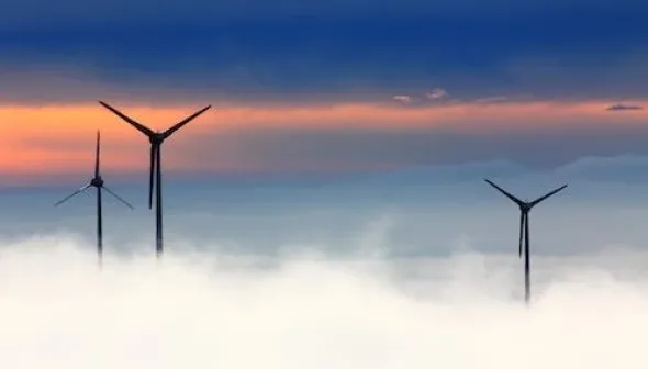 Three wind turbines can be seen peeking out above wispy white clouds in front of an orange-streaked dark blue sky.