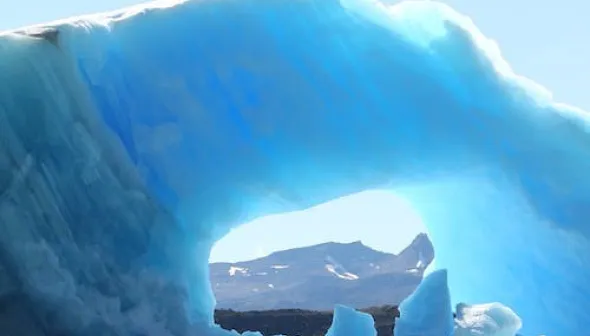 Un énorme morceau de glace flottant dans l'eau remplit le cadre.  Un trou au centre de la glace nous permet de voir une montagne au loin derrière.
