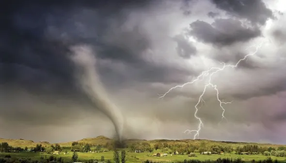 A tornado funnel is shown touching down onto green hilly farmland below a dark stormy sky with lightning bolts.  Various homes and barns can be seen in the distance.