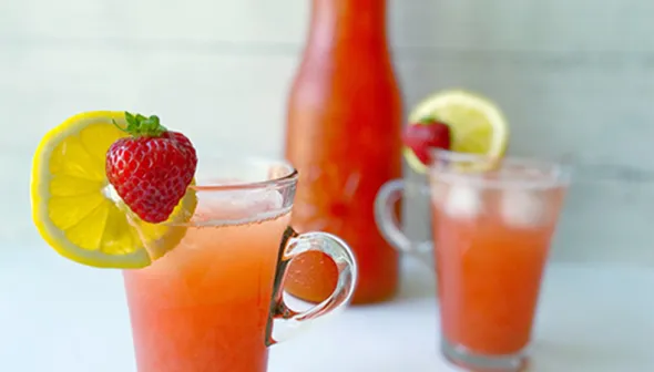 A tall glass of bright pink lemonade stands on a counter in the foreground. In the background, another glass and a carafe filled with lemonade are visible. Lemon wedges and strawberries decorate the rims of the glasses.
