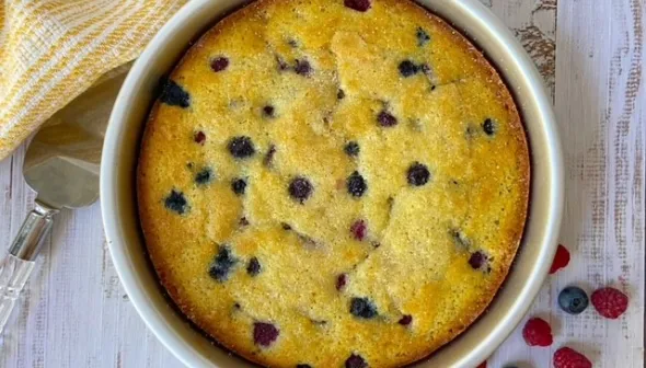 An aerial view shows a Berry Buttermilk cake in a round pan, sitting on top of a faded wooden surface. Assorted berries are sprinkled next to the pan.