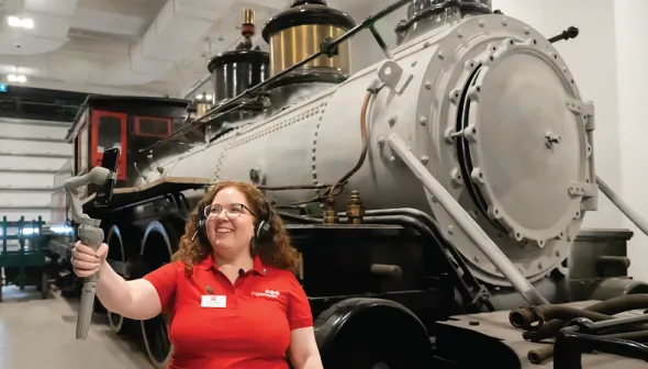  Une employée de musée se tient devant une grande locomotive à vapeur grise et noire. Elle porte un écouteur et sourit à l'écran d'un téléphone intelligent qu'elle tient par un cardan dans son bras tendu.