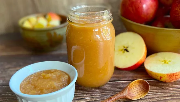 A large jar and a small bowl of applesauce sit on top of a wooden surface. Whole and chopped apples are visible in the background.