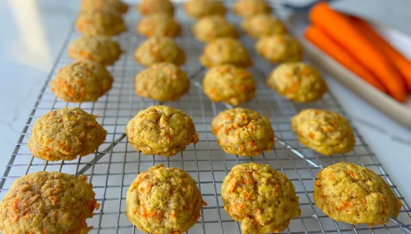 Rows of carrot cookies are lined up on a baking rack, set against a white background. A dish of orange carrots is visible in the background.