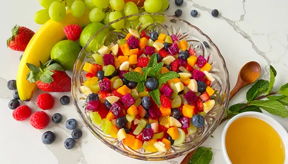 An aerial view depicts a colourful bowl of fruit salad on a marble countertop. An assortment of fruits, a spoon and a small bowl of dressing are next to the salad.