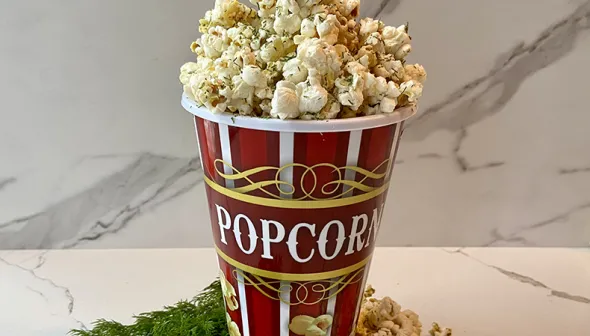 A large bowl of dill popcorn rests against a marbled wall. Fresh dill is seen at the front with a small bowl of corn kernels.  