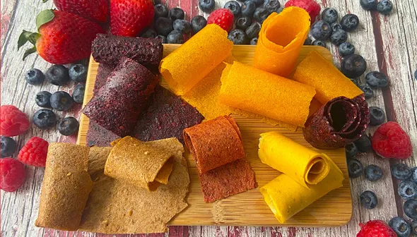 A number of fruit leather roll-ups sit on a wooden surface, with mixed berries sprinkled around them.