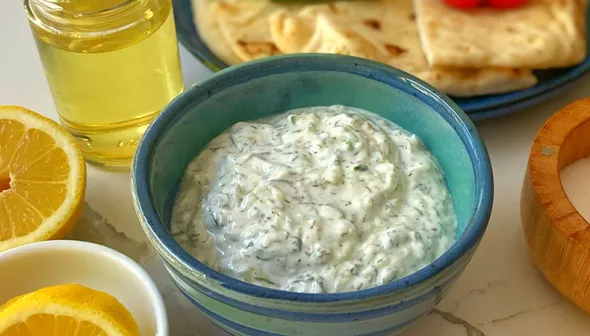 A blue bowl of dip sits on a white tabletop. Lemon wedges, olive oil and a salt bowl are visible around the dip. A blue plate with vegetables and pita wedges is visible in the background.  