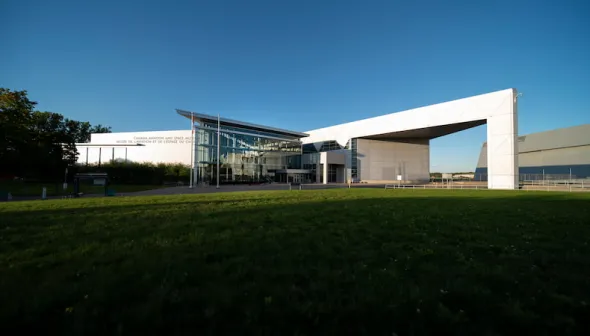La façade angulaire d’un grand bâtiment blanc, avec les mots « Musée de l'aviation et de l'espace du Canada » visibles sur son côté gauche. De l’herbe verte est visible en avant-plan et un ciel bleu et dégagé en arrière-plan.