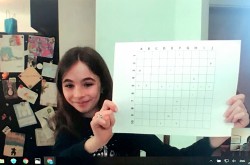 Image of a video chat taken from a computer screen. A girl appears onscreen, smiling and holding up a grid covered with coloured markings.