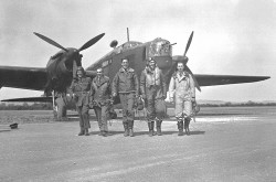 Cinq hommes, vêtus de leur équipement de vol, se dirigent vers la caméra avec un bombardier, vu de l’avant et de la gauche, à l’arrière-plan.