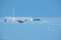 A barren, snowy landscape is set against a blue sky. A research facility for Environment and Climate Change Canada can be seen, with its lights glowing.