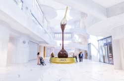 A wide shot of a gallery inside the Lindt Home of Chocolate, showing white walls, white marble floors and a massive chocolate fountain. A few people are standing and sitting around the fountain, looking up at it.