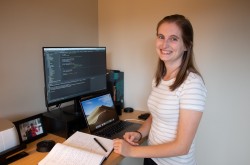 Une jeune femme souriante est debout à son poste de travail; devant elle se trouvent un ordinateur, un écran, un cahier de notes ouvert et un stylo.
