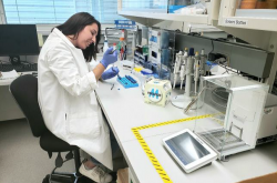 Une jeune femme portant une blouse de laboratoire blanche et des gants de caoutchouc bleus travaille assise dans un laboratoire; plusieurs outils sont disposés devant elle.