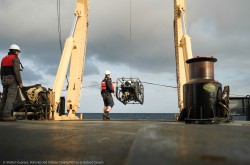 Une grue à l'arrière d'un navire abaisse un robot d'exploration des fonds marins pendant que deux ouvriers observent