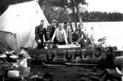 Une image en noir et blanc présente un groupe de sept hommes debout derrière un gros morceau de bois sur lequel sont peintes les lettres d’appel « G-CAAC ». Ils se tiennent près d’un lac. Derrière eux se trouvent une tente blanche et un petit groupe de trois arbres. Devant eux, du côté gauche de la photo, on voit une pile d’équipement de camping et de cuisson. 