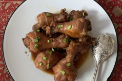 A plate of medium-brown soy sauce chicken drumsticks. Green onion pieces are sprinkled on top, and a silver pair of tongs rests on the plate. A red floral placemat can be seen under the white plate. 