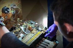 Over-the-shoulder shot of curator and conservator peering under the hood of the Electronic Sackbut synthesizer. Internal wiring and circuitry behind keyboard is exposed. 