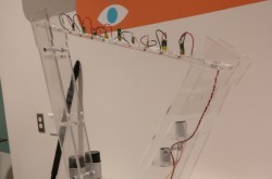 A clear, plastic harp-shaped frame has a series of colourful wires coming out of the top. The harp sits on a countertop against a wall; a colourful mural can be seen in the background.