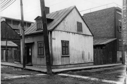 Une image noir et blanc montre une maison en bois dans un quartier ouvrier, au début des années 1900.]