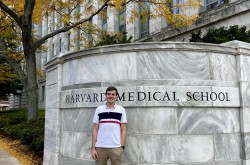 Award winner Jackson Weir stands next to a large marble wall with a sign that reads, "Harvard Medical School." A tree and the facade of a building are visible in the background.