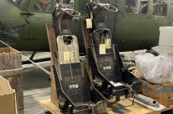 A pair of bare metal ejection seats with no cushions or padding are mounted on a plywood base, placed beside a few cardboard boxes. Behind the seats, a camouflage-green helicopter is partially visible.