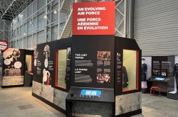 Several exhibition display cases, panels, and screens in a large, brightly-lit air hangar-style room with a high ceiling and tall, exposed metal walls.